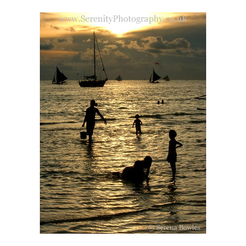 Silhouetted boats, and figures of children at play, against the setting sun reflected in the water.